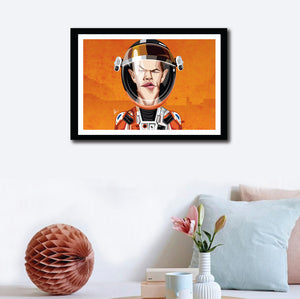 Matt Damon in his Caricature Art Form by Prasad Bhat. Image shows wall decor of the framed art poster of Martian avatar by Matt looking straight forward with his sleek eyes . He is wearing the astronaut's suit and helmet against a predominantly orange background.